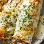 Baked chicken burritos topped with creamy green chile sauce on a plate.