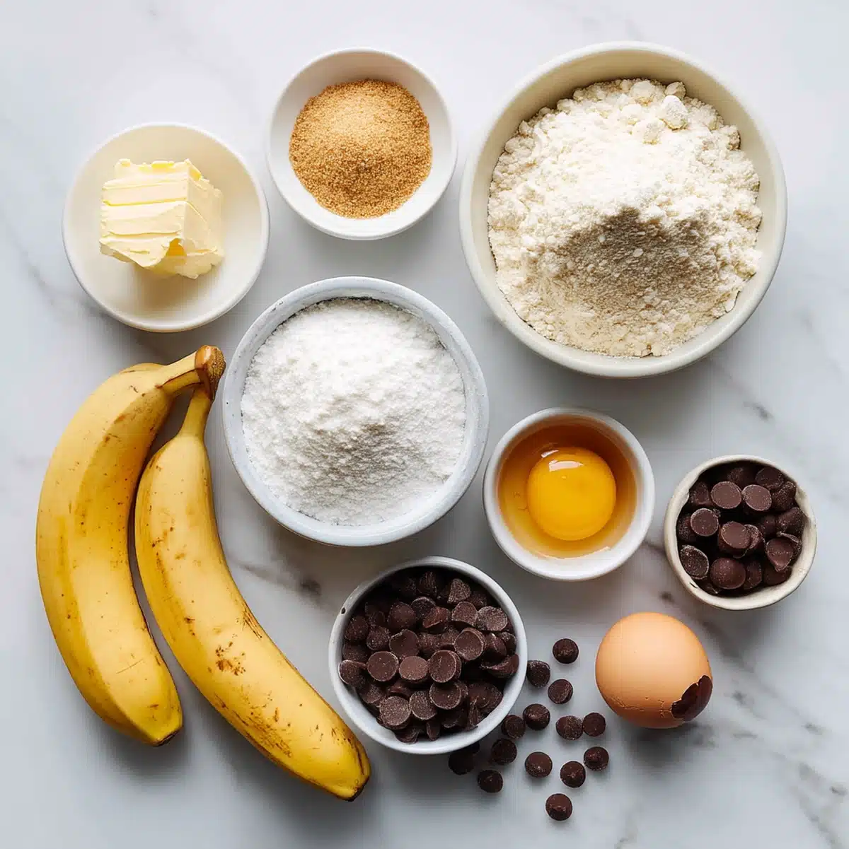 Flat lay of brown-speckled ripe bananas, unbleached flour, and mini chocolate morsels on a wooden surface